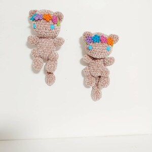 K&ouml;nnte beinhalten: Zwei geh&auml;kelte Pl&uuml;schkatzenspielzeuge mit langen Schw&auml;nzen. Beide Katzen tragen Blumenkr&auml;nze mit rosa, lila, orangefarbenen und blauen Blumen. Die Katzen haben blaue Augen.