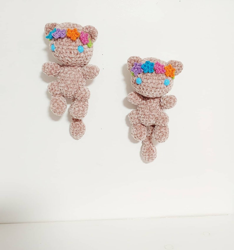 K&ouml;nnte beinhalten: Zwei geh&auml;kelte Pl&uuml;schkatzenspielzeuge mit langen Schw&auml;nzen. Beide Katzen tragen Blumenkr&auml;nze mit rosa, lila, orangefarbenen und blauen Blumen. Die Katzen haben blaue Augen.
