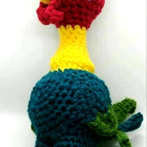 May include: A handmade crochet rooster toy. The rooster has a red head and comb, a yellow beak, and a white eye with a black pupil. The body is teal, with green tail feathers and yellow feet. A whimsical, colorful creation.