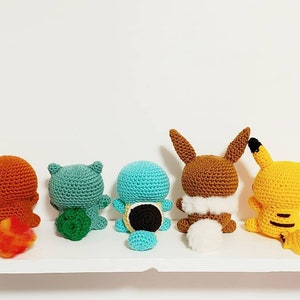 K&ouml;nnte beinhalten: F&uuml;nf geh&auml;kelte Pok&eacute;mon-Charaktere, darunter Glumanda, Bisasam, Schiggy, Evoli und Pikachu, stehen auf einem wei&szlig;en Regal. Die Figuren sind mit dem R&uuml;cken zum Betrachter ausgerichtet und zeigen ihren R&uuml;cken.
