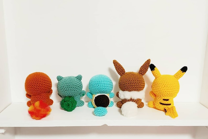 K&ouml;nnte beinhalten: F&uuml;nf geh&auml;kelte Pok&eacute;mon-Charaktere, darunter Glumanda, Bisasam, Schiggy, Evoli und Pikachu, stehen auf einem wei&szlig;en Regal. Die Figuren sind mit dem R&uuml;cken zum Betrachter ausgerichtet und zeigen ihren R&uuml;cken.