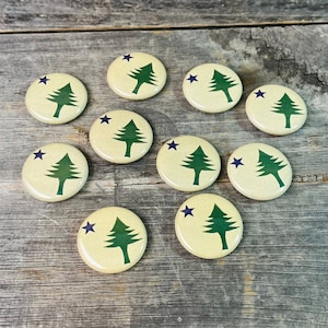 May include: A collection of ten round buttons with a cream-colored background. Each button features a green pine tree and a dark blue star. The buttons are arranged on a weathered wooden surface.