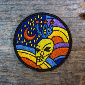 May include: A colorful embroidered patch featuring a skull with a cactus growing out of its head. The background is a swirling pattern of blue, yellow, orange, and purple.