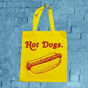 Puede incluir: Bolso tote amarillo con texto rojo "Hot Dogs" y una ilustración de un perrito caliente. El bolso tiene asas amarillas y está sobre una pared de ladrillos azules.