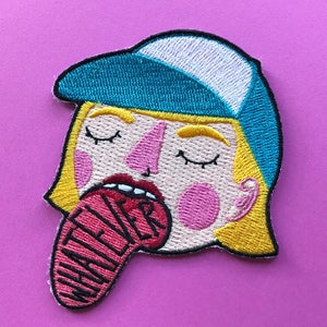 May include: Embroidered iron-on patch featuring a cartoon character with blonde hair, blue eyes, and a blue baseball cap. The character is sticking out their tongue with the word "Whatever" written on it.