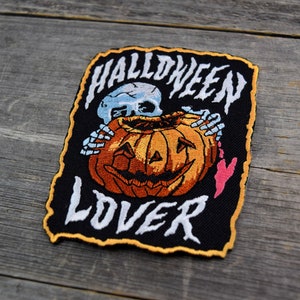 K&ouml;nnte beinhalten: Schwarzer gestickter Patch mit einem Skelett, das einen K&uuml;rbis h&auml;lt. Der Text "HALLOWEEN LOVER" ist in Wei&szlig; auf den Patch gestickt.