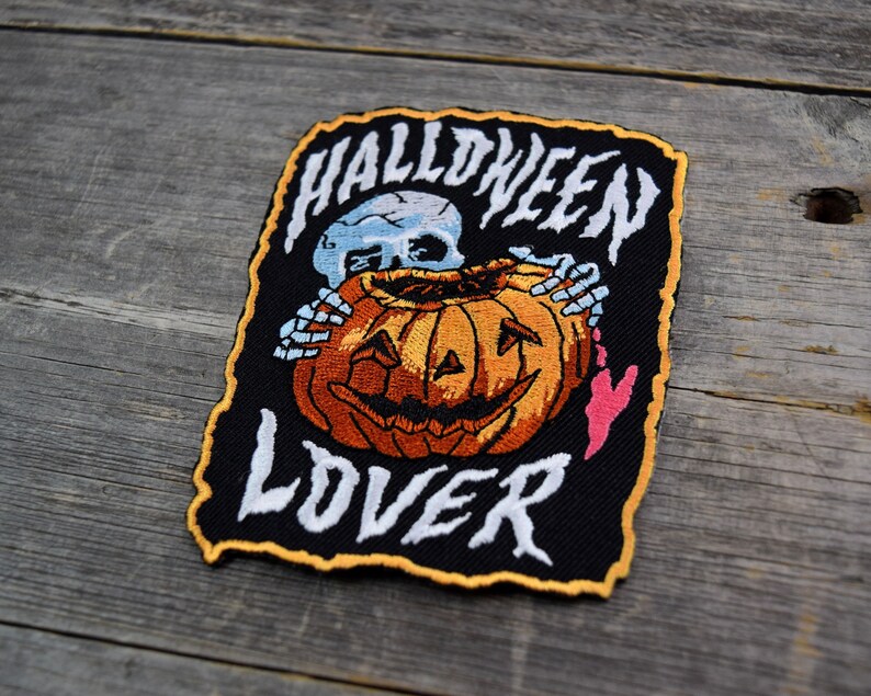 K&ouml;nnte beinhalten: Schwarzer gestickter Patch mit einem Skelett, das einen K&uuml;rbis h&auml;lt. Der Text "HALLOWEEN LOVER" ist in Wei&szlig; auf den Patch gestickt.