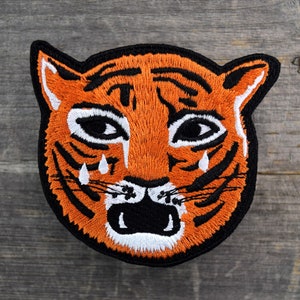 May include: Embroidered iron-on patch of a tiger's face with black stripes and a sad expression. The tiger has white teardrops coming from its eyes.