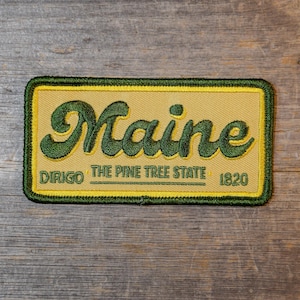 Puede incluir: Parche rectangular con fondo beige y borde verde. La palabra "Maine" está bordada en letras verdes grandes y cursivas. Debajo, se lee "DIRIGO THE PINE TREE STATE 1820" en letras verdes más pequeñas.