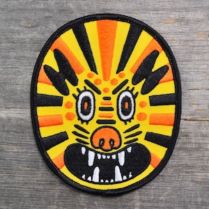May include: Oval-shaped embroidered patch featuring a stylized tiger face. The design includes a yellow background with orange and black stripes, black accents, and a detailed face with white teeth.