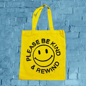 Op de afbeelding: Een heldergele draagtas met bijpassende handvatten. De tas heeft een zwarte smiley en de tekst "PLEASE BE KIND & REWIND" in een verweerde lettertype. De tas is gemaakt van een stevige stof.