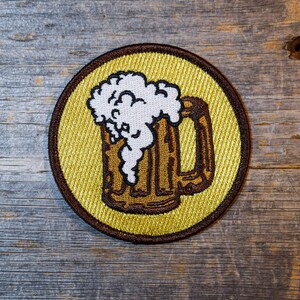 May include: A brown and white embroidered patch of a beer mug with foam on top, on a yellow background.