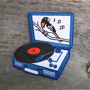 May include: A blue and white embroidered patch of a vintage record player with a black record and a red label. A bird is singing in the record player's lid, with musical notes above it.