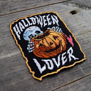 K&ouml;nnte beinhalten: Schwarzer gestickter Patch mit einem wei&szlig;en Skelett, das einen geschnitzten orangefarbenen K&uuml;rbis mit einem gruseligen Gesicht h&auml;lt. Der Text "HALLOWEEN LOVER" ist in Wei&szlig; auf dem Patch gestickt.