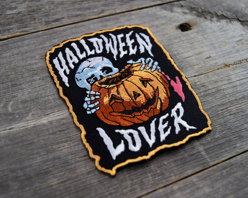K&ouml;nnte beinhalten: Schwarzer gestickter Patch mit einem wei&szlig;en Skelett, das einen geschnitzten orangefarbenen K&uuml;rbis mit einem gruseligen Gesicht h&auml;lt. Der Text "HALLOWEEN LOVER" ist in Wei&szlig; auf dem Patch gestickt.