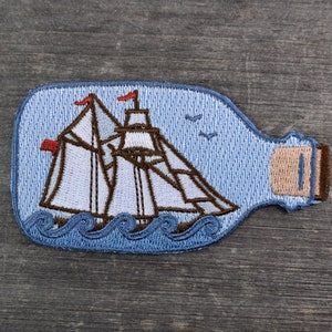 May include: A blue embroidered patch of a ship in a bottle. The ship has white sails and brown masts. The bottle is blue with a brown cork.