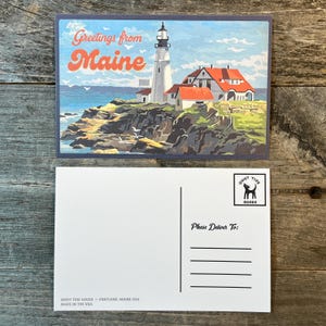 May include: A postcard featuring a colorful illustration of a lighthouse on a rocky coast with the text "Greetings from Maine." The back of the postcard has lines for an address and the text "Please Deliver To:" and the Quiet Tide Goods logo.