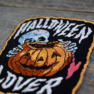 K&ouml;nnte beinhalten: Ein schwarzer gestickter Aufn&auml;her mit einem Skelett, das einen K&uuml;rbis h&auml;lt. Der Aufn&auml;her tr&auml;gt die Aufschrift "Halloween Lover" in wei&szlig;en Buchstaben.