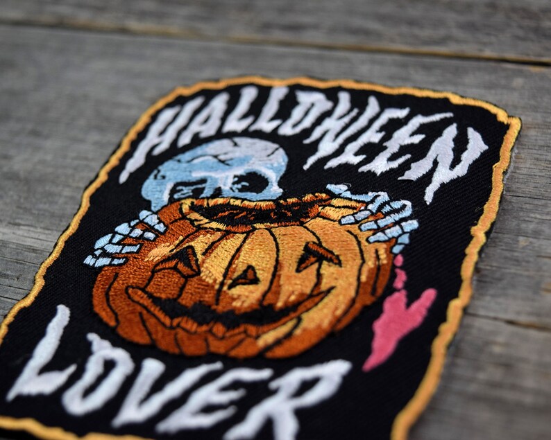 K&ouml;nnte beinhalten: Ein schwarzer gestickter Aufn&auml;her mit einem Skelett, das einen K&uuml;rbis h&auml;lt. Der Aufn&auml;her tr&auml;gt die Aufschrift "Halloween Lover" in wei&szlig;en Buchstaben.