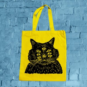 May include: A bright yellow tote bag featuring a black illustration of a cat with six eyes. The cat's fur is detailed with fine lines, and the bag has matching yellow handles.