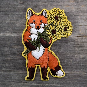 May include: Embroidered patch featuring a cartoon fox holding a bouquet of sunflowers. The fox has orange and white fur with brown accents. The sunflowers are yellow with brown centers and green stems. The patch has a yellow border.