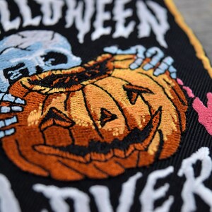 K&ouml;nnte beinhalten: Gestickter Aufn&auml;her mit schwarzem Hintergrund, einem wei&szlig;en Skelett, das einen geschnitzten orangefarbenen K&uuml;rbis mit einem zahnigen Grinsen h&auml;lt, und dem Text "Halloween Forever" in wei&szlig;.