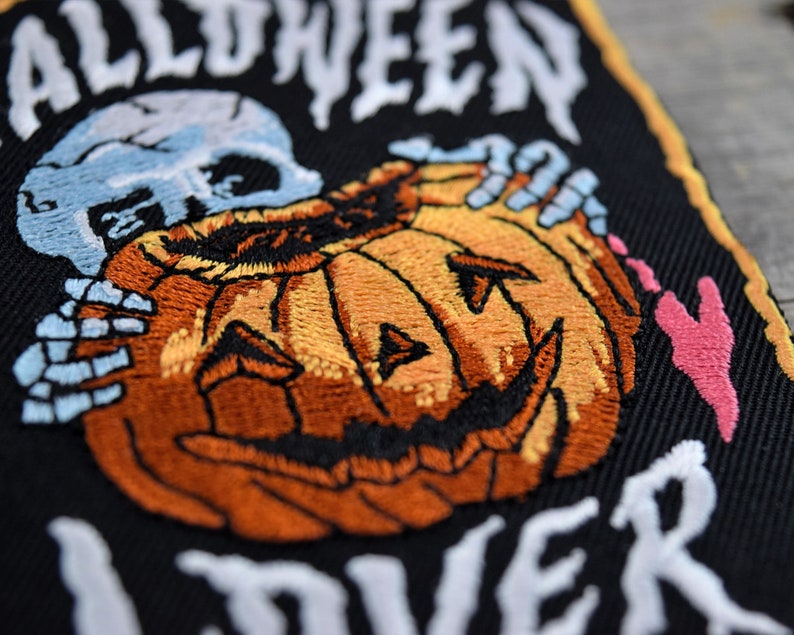 K&ouml;nnte beinhalten: Gestickter Aufn&auml;her mit schwarzem Hintergrund, einem wei&szlig;en Skelett, das einen geschnitzten orangefarbenen K&uuml;rbis mit einem zahnigen Grinsen h&auml;lt, und dem Text "Halloween Forever" in wei&szlig;.