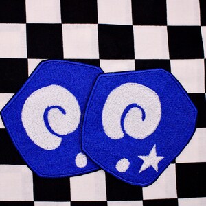 May include: Two blue, hexagon-shaped embroidered patches with white spiral designs, a white star, and white dots. The patches are set against a black and white checkered background.