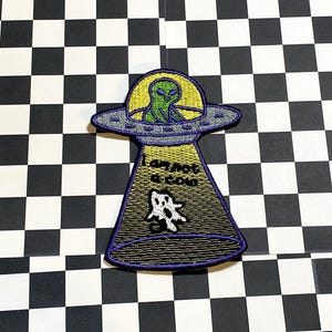 May include: Embroidered patch featuring a green alien in a UFO. The UFO is purple and yellow, with the text "I am not a cow" and a cow graphic. The patch is on a black and white checkered background.