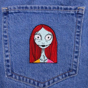 May include: A denim jacket pocket featuring an embroidered patch of Sally from The Nightmare Before Christmas. The patch shows Sally's face with red hair, a blue face, and a stitched mouth. The pocket is a medium blue color.