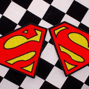 May include: Two embroidered patches featuring the Superman logo. The patches are red with a yellow "S" inside a shield shape, outlined in black. The background is a black and white checkered pattern.