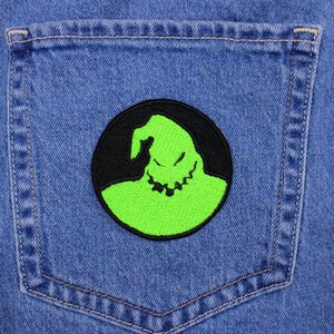 May include: A round patch featuring a neon green character with a jagged smile and a black outline, set against a black background. The patch is affixed to the back pocket of a blue denim jacket.