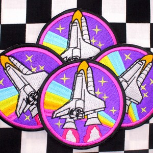 May include: Five embroidered patches featuring a space shuttle design. Each patch has a purple background with a pink border, a white shuttle, and yellow, blue, and orange accents. The patches are on a black and white checkered background.