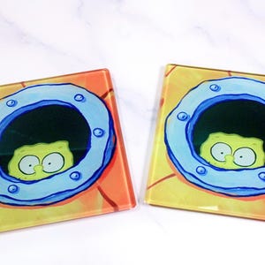 May include: Two square glass coasters featuring a graphic print of a yellow sponge character peering through a blue circular porthole. The background of the coasters is a warm orange and yellow gradient, mimicking the appearance of a submarine wall.