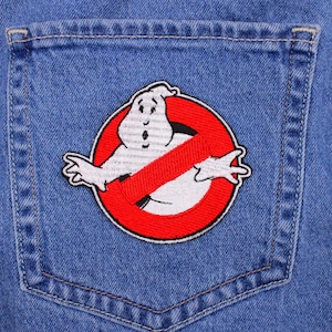 May include: A blue denim jacket pocket featuring a Ghostbusters patch. The patch is a white ghost inside a red circle with a red slash through it. The patch is embroidered with black and white details.