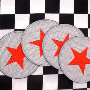 May include: Four gray circular patches with a red star design. The patches are embroidered with a textured pattern and a black border. The background is a black and white checkered pattern.