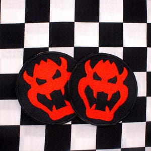 May include: Two round black patches with a red Bowser design are displayed on a black and white checkered background. The patches feature a detailed, embroidered image of Bowser's face.
