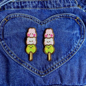 May include: Close-up of a denim overall pocket with a heart-shaped design. Two embroidered patches of kawaii dango, a Japanese dessert, are sewn onto the pocket. Each patch features a pink, white, and green dango stack.