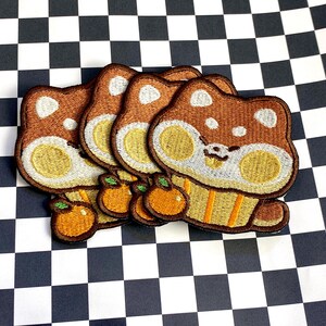 May include: A stack of embroidered patches featuring a cartoon bear design. The patches are brown, tan, and orange, with a white accent on the head and a small orange fruit. The patches are on a black and white checkered background.