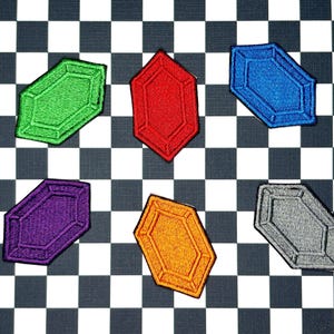 May include: Six colorful, embroidered patches in the shape of stylized gems. The patches are green, red, blue, purple, orange, and gray. The patches are arranged on a black and white checkered background.