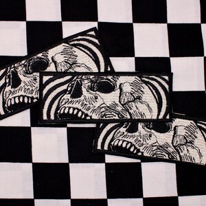 May include: Four rectangular patches with a skull design. The skulls are black and white, with a detailed illustration of the skull's features. The patches are on a black and white checkered background.