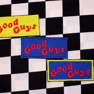 May include: Three embroidered patches with the text "Good Guys" in red lettering. One patch is yellow with a black border, and two are blue with yellow borders. The patches are on a black and white checkered background.