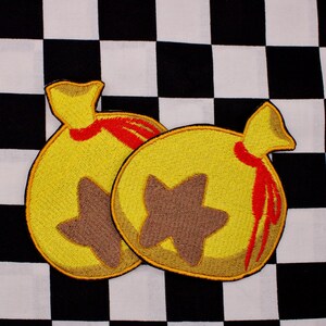 May include: Two yellow embroidered money bag patches with brown star designs and red accents. The patches are on a black and white checkered background. The patches are likely for a video game.