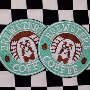 May include: Two round embroidered patches with the text "BREWSTER'S COFFEE" in white on a teal background. The patches feature a cartoon character with a mustache, glasses, and a bow tie. Coffee beans are also included in the design. The background is a black and white checkered pattern.