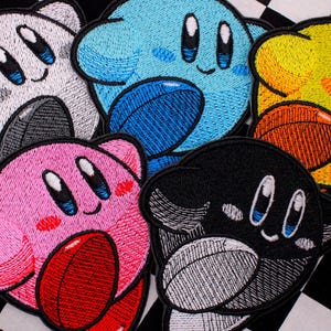 May include: Embroidered patches featuring a cartoon character in various colors: white, blue, yellow, pink, and black. Each patch has a smiling face and holds a football-shaped object. The patches are on a black and white checkered background.