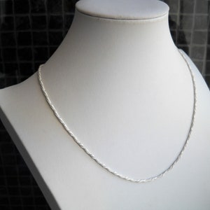 May include: A silver-colored rope chain necklace displayed on a white mannequin. The chain has a twisted design and is draped around the mannequin's neck. The background is a dark, textured surface.