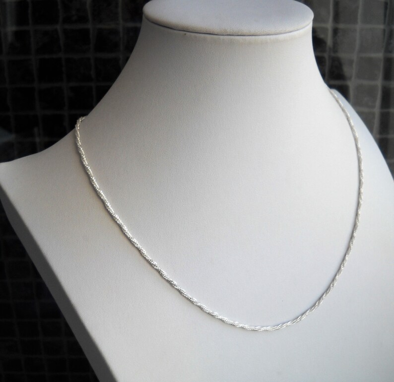 May include: A silver-colored rope chain necklace displayed on a white mannequin. The chain has a twisted design and is draped around the mannequin's neck. The background is a dark, textured surface.