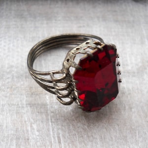 Pode incluir: Um anel de prata com uma grande gema vermelha facetada engastada em uma configuração decorativa de trabalho aberto.