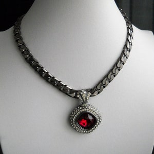 Pode incluir: Um colar em tom de metal com um grande pingente vermelho rubi quadrado. O pingente é cercado por pequenos cristais transparentes. A corrente é um design espesso e ligado. O colar é exibido em um manequim branco.