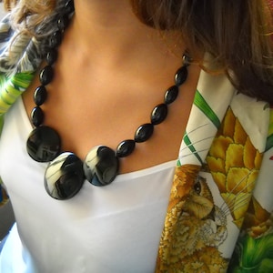 Peut inclure: Un collier noir composé de perles brillantes, rondes et ovales. Le collier est porté sur un haut blanc, avec une écharpe colorée ornée d'un motif de lapin et de plantes.